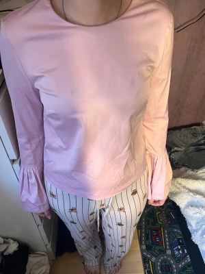 Ljusrosa blus med volangärmar - Söt ljusrosa blus i glansigt material med rund halsringning och långa ärmar som avslutas med volangdetalj vid ärmsluten. Perfekt för en trendig och feminin look. Enkel att matcha med både jeans och kjol.