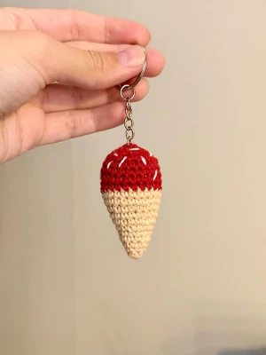 Handvirkad glass-strut  - Säljer en handgjord virkad nyckelring formad som en glass-strut med röd och beige färg. Glassens topp är dekorerad med vita detaljer som ser ut som strössel. Perfekt att hänga på väskan eller nyckelknippan.