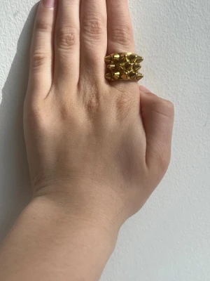 Guldig statementring med nitar - Cool och chunky ring i guldigt metallmaterial med flera små nitar på ovansidan. Ringen har en bred design och ger en edgy vibe till din stil. Perfekt för dig som gillar att sticka ut med accessoarer.