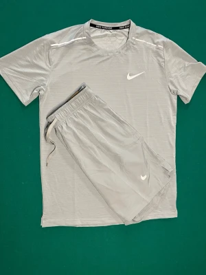 Nike Miller set - Säljer ett grått träningsset från Nike med t-shirt och shorts. Båda plaggen har den klassiska Nike-loggan i vitt. Materialet är lätt och andas, perfekt för löpning eller gym. Shortsen har resårmidja med dragsko för skön passform.
