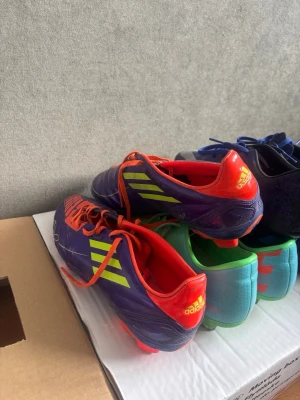 Adidas lila fotbollsskor med neonränder - Säljer ett par snygga Adidas fotbollsskor i lila med neonlimegröna ränder och detaljer. Skorna har orange snörning och insida samt dobbar för spel på gräs. Perfekta för dig som vill sticka ut på planen och få bra grepp i spelet.