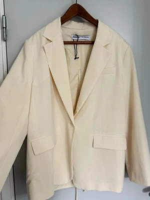 Krämvit kavaj från Bershka - Stilren krämvit kavaj från Bershka med klassisk krage, två framfickor med lock och en bröstficka. Kavajen har en unik detalj med knytband i ryggen och är tillverkad i ett mjukt tyg som ger en snygg passform. Perfekt för dig som vill ha en modern twist på din outfit. Liten fläck på ryggen syns inte mycket och går säkert att tvätta bort 