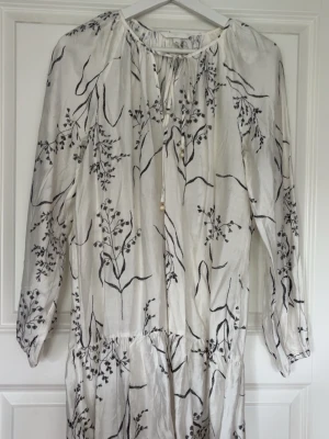 Mini klänning - Mini klänning från H&M i vit med svart blommigt mönster. Klänningen har långa ärmar, knytning vid halsen och är tillverkad i ett lätt och luftigt material som känns skönt mot huden. Perfekt för en avslappnad och stilren look.