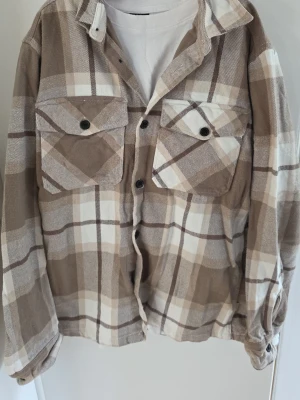 Beige och vit rutig overshirt från jack and jones - Overshirt i beige, vitt och brunt rutmönster med två stora bröstfickor och svarta knappar. Skön och avslappnad passform, perfekt att bära över en t-shirt. Tjockare bomullsmaterial som ger en mjuk känsla.
