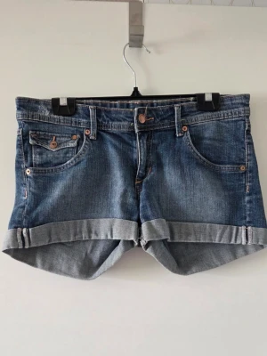 Blå jeansshorts - Snygga blå jeansshorts med uppvikta benslut och klassiska fem fickor. Shortsen har knapp- och dragkedjestängning samt detaljer i kopparfärg. Perfekta för varma dagar och enkla att matcha med olika stilar.
