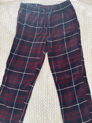 Rutiga pyjamasbyxor i rött och blått - Mysiga pyjamasbyxor med rött, mörkblått och vitt rutmönster. Byxorna har resår i midjan och dragsko för skön passform. Perfekta för chillkvällar hemma. Materialet känns mjukt och bekvämt mot huden.