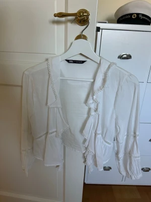 Vit boho-blus med volang från Zara - Säljer en luftig vit blus från Zara med volangkant och dekorativa spetsdetaljer. Blusen har lång ärm med resår vid ärmslut och en avslappnad, bohemisk look. Perfekt att styla med jeans eller kjol för en trendig outfit.
