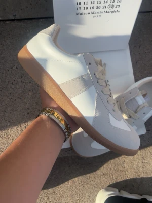 Maison Margiela vita sneakers - Maison Margiela sneakers i vitt och ljusgrått med beige mockadetaljer och klassisk gummisula. Skorna har en stilren retrodesign med vita läderpaneler, beige snören och låg silhuett. Perfekta för dig som gillar minimalistisk och trendig stil.