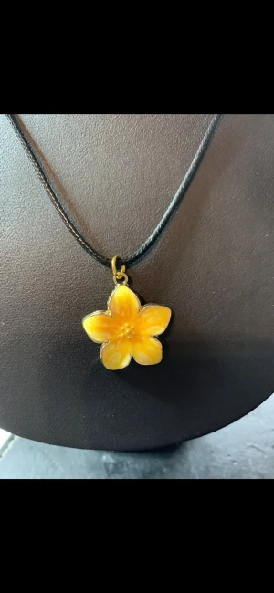 Halsband med gul blomma i emalj - Snyggt halsband med ett hängsmycke i form av en gul blomma. Blomman har fem kronblad och är gjord i emalj med fina detaljer. Hänget sitter på ett svart, tunt rem som ger en stilren look. Perfekt för dig som vill ha något färgglatt och unikt.