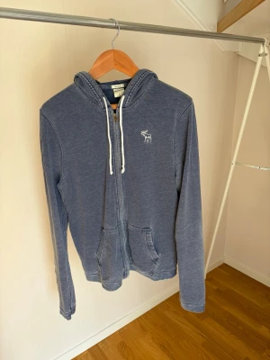 Blå zip hoodie med vit logga - Säljer en blå hoodie med dragkedja och huva, vita snören och en liten vit logga på bröstet. Hoodien har två fickor framtill och är tillverkad i mjukt bomullsmaterial. Perfekt för en avslappnad och chill stil.