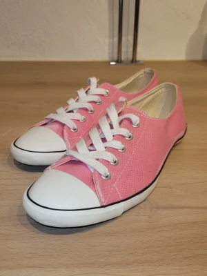 Rosa låga tygsneakers med vita detaljer - Säljer ett par klassiska låga sneakers i rosa tyg med vita gummisulor och tåhättor. Skorna har vita skosnören och silverfärgade öljetter. Perfekta för dig som vill ha en färgklick till din outfit. Helt oanvända. 