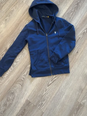 Blå hoodie från Polo Ralph Lauren - Mörkblå hoodie med dragkedja från Polo Ralph Lauren. Klassisk design med huva, dragsnören och fickor framtill. Ikonisk gul broderad logga på bröstet. Tillverkad i mjukt bomullsmaterial som är skönt att bära.
