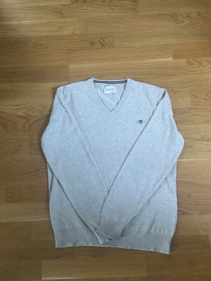 Ljusgrå/beige V-ringad tröja från Gant - Ljusgrå V-ringad tröja från Gant med broderad logga på bröstet. Tröjan har långa ärmar, ribbade muddar och är gjord i ett tunt, mjukt material. Enkel och stilren design som är lätt att matcha. Cirka 2 centimer lagning vid nedre mudd (se bild).