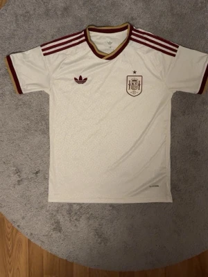 Vit Spanien fotbollströja Adidas - Säljer en vit Spanien fotbollströja från Adidas med vinröda detaljer och guldbrun krage. Tröjan har tre klassiska Adidas-ränder på axlarna, Spanien-emblem på bröstet och Adidas-logga. Tillverkad i lätt och ventilerande material, perfekt för träning eller match.