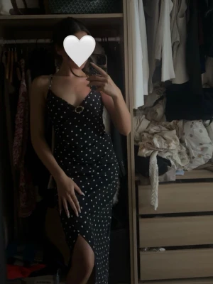 Svart prickig klänning med slits - Snygg svart klänning med vita prickar och smala axelband. Klänningen har en v-ringad urringning med dekorativ ringdetalj och en slits framtill som ger en trendig look. Perfekt för dig som vill sticka ut lite extra.