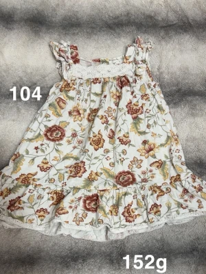 Blommig volangklänning - Söt kortklänning med volangkant och blommigt mönster i beige, gul och röd. Klänningen har breda axelband med volangdetaljer och spets upptill. 🌸Fint använt skick. Storlek 104