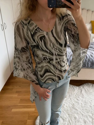 Mönstrad långärmad blus i beige och svart - Säljer en snygg långärmad blus med vågigt mönster i olika nyanser av grönt och vitt. Blusen har vida ärmar och är gjord i ett tunt, lätt material som känns luftigt. Perfekt att styla med jeans eller kjol för en trendig look. Står storlek M, men passar mer som en S. 