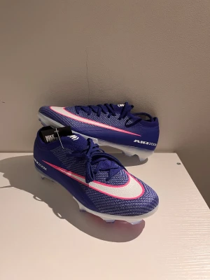 Nike Air elite  Zoom Mercurial blå fotbollsskor - Snygga Nike Air Zoom Mercurial fotbollsskor i blått med vita och rosa detaljer. Skorna har en strömlinjeformad design, syntetiskt material och snörning för optimal passform. Perfekta för snabba vändningar på planen och har en lätt yttersula med dobbar för bra grepp.