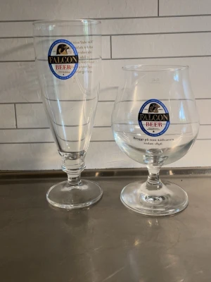 Falcon Beer ölglas 80kr st - Två olika ölglas från Falcon Beer, båda i genomskinligt glas med Falcon-logga framtill. Ett glas är högt och smalt, det andra är lägre och rundare med bred fot. Perfekta för att njuta av öl med stil.