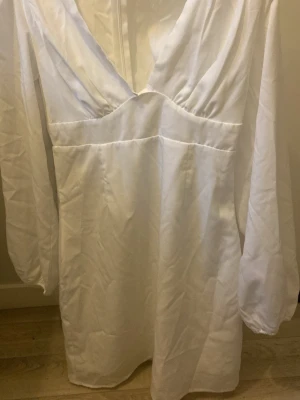 Vit omlottklänning med v-ringning - Stilren vit klänning med omlottdesign och djup v-ringning. Klänningen har långa ärmar och är tillverkad i ett lätt, glansigt material som ger en elegant känsla. Perfekt för dig som gillar minimalistisk och chic stil.