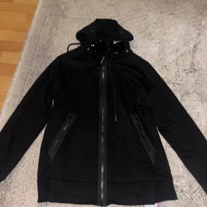 Cp Company hoodie  - Svart vindjacka med coola inbyggda glasögon i huvan och dragkedjor på fickorna. Jackan har lång ärm, hel dragkedja framtill och snörning vid huvan. Perfekt för dig som gillar streetwear och vill sticka ut med en unik detalj.