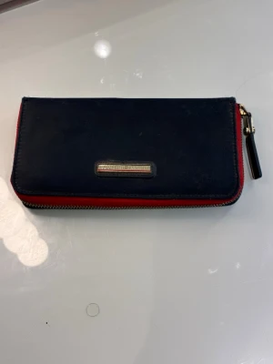  Marinblå Tommy Hilfiger plånbok med röd kant - Stilren marinblå plånbok med röd kant och guldfärgad dragkedja. Plånboken har en metallbricka framtill och dragkedjan har ett dekorativt band. Tillverkad i syntet och har en rektangulär form som rymmer kort och sedlar. Storlek 19cm*10cm. Den ser fräsch ut på både ut och insidan, men såg när jag inspekterade den invändigt att den släppt lite i sömnen på två ställen så att det blivit två hål (se sista två bilderna ).Det är dock inget som påverkar varesig utseende eller funktion. 