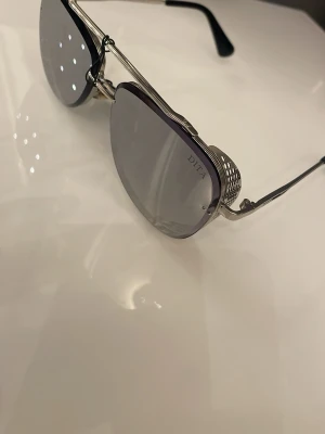 Silvriga solglasögon från Dita - Unika solglasögon från Dita med silvriga metallbågar och gråtonade glas. Sidorna har coola mesh-detaljer som ger en futuristisk vibe. Perfekta för dig som vill sticka ut och addera en edgy touch till din stil.
