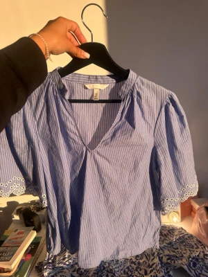 Blåvit randig blus från H&M - Söt blå blus med vita ränder från H&M. Blusen har korta puffärmar med broderade spetsdetaljer vid ärmsluten och en djup v-ringning. Tillverkad i ett lätt och luftigt material, perfekt för varma dagar. Fläck på bild tre!!!!
