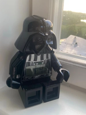 LEGO Star Wars Darth Vader väckarklocka - Cool digital väckarklocka formad som Darth Vader i LEGO-stil. Klockan är svart med vita och grå detaljer på bröstet och har en tydlig digital display på magen. Perfekt för Star Wars-fans som vill ha något unikt och nördigt på nattduksbordet.