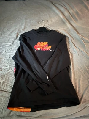 Svart långärmad Jumpman topp - Säljer en svart långärmad topp med färgglatt 'Jumpman'-tryck framtill. Tröjan har rund halsringning och coola detaljer i rött, gult och orange på trycket. Perfekt för dig som gillar streetwear och sportig stil. Priset är Förhandlingsbart.
