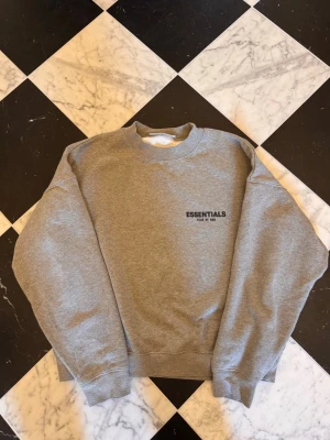 Grå sweatshirt från Essentials - Säljer en stilren grå sweatshirt från Essentials Fear of God. Tröjan har rund halsringning, ribbade muddar och diskret svart logga på bröstet. Perfekt för en clean och avslappnad look.
