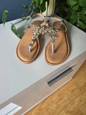 Silvriga sandaler med strassdetaljer - Snygga sandaler med silvriga remmar och dekorativa strass och pärlor på ovandelen. Sandalerna har en låg klack och öppen tå, samt en mjuk, beige innersula för extra komfort. Perfekta för att ge din outfit lite extra bling.                 OBS!!! Storlek 40 men det gick bara att välja 39