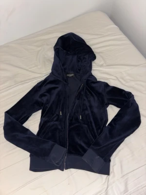 Mörkblå hoodie Juicy Couture - Mjuk och lyxig mörkblå hoodie från Juicy Couture i velourmaterial. Dragkedja framtill, två fickor och justerbar huva med snören. Ribbstickade muddar vid ärmslut och nederkant. Perfekt för en chill och trendig look.