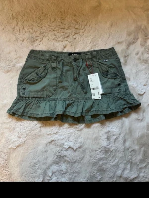 Grön volangkjol från Urban outfitters  - Snygg grön kortkjol från Urban outfitters med volangkant och dekorativa fickor framtill. Kjolen har knytband i midjan och detaljer med knappar. Perfekt för dig som vill ha en avslappnad men trendig look.