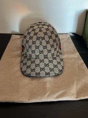 Gucci keps beige  - Snygg keps från Gucci i beige med klassiskt GG-monogram över hela kepsen. Kepsen har böjd skärm och en röd-blå rand på sidan. Tillverkad i canvasmaterial och levereras med original dustbag. Perfekt för dig som vill ha en exklusiv och trendig accessoar.