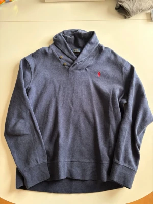 Blå tröja från Polo Ralph Lauren - Blå tröja från Polo Ralph Lauren med hög krage och två knappar vid halsen. Tröjan har lång ärm och den klassiska röda loggan broderad på bröstet. Perfekt för dig som gillar stilren och avslappnad look.