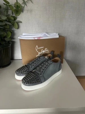  Christian Louboutin sneakers - Snygga gråa sneakers från Christian Louboutin med klassiska röda sulor och coola silverfärgade nitar på tån. Skorna är låga och har en ovandel i mocka med vita gummisulor. Perfekta för dig som vill sticka ut med en lyxig och edgy stil. Box och dustbag medföljer, 42. Boxen är lite sönder. Skorna är blå/grå typ