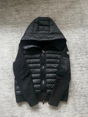 Svart Moncler Cardigan - Säljer en svart cardigan från Moncler med stickade ärmar och quiltad kropp. Jackan har en stor huva, dragkedja framtill och en liten ficka med Moncler-logga på ärmen. Perfekt för kalla vinterdagar och riktigt snygg streetstil. Den svarta färgen på de stickade området har blekts lite grann av solen. 