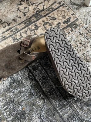 Bruna Birkenstock Boston tofflor mocka - Säljer ett par klassiska Birkenstock Boston tofflor i brun mocka med justerbart spänne i metall. Tofflorna har en anatomiskt utformad fotbädd och mönstrad gummisula
