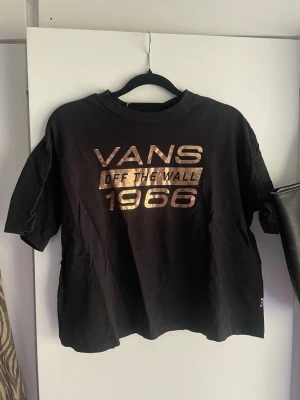 Svart Vans t-shirt med guldtryck - Cool svart t-shirt från Vans med metallic guldtext 'VANS OFF THE WALL 1966' på bröstet. Klassisk passform med rund hals och korta ärmar. Storlek XS