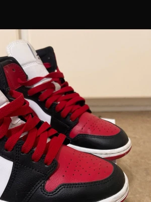 Air Jordan 1 Mid röd svart vit - Säljer ett par Air Jordan 1 Mid i klassisk röd, svart och vit färg. Skorna har röd swoosh, röda skosnören och svart läderdetaljer. Ikonisk Jordan-logga på sidan och vit mellansula. Perfekta för dig som gillar streetwear och sneakerskultur.