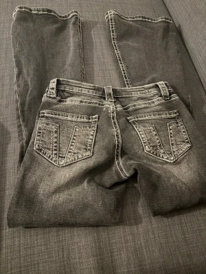 Grå bootcut jeans  - Säljer ett par grå bootcut jeans med tydliga kontrastsömmar och klassiska fem fickor. Jeansen har en lätt tvättad look och utsvängda ben. Perfekta för dig som gillar retrostil och vill ha ett par jeans som sticker ut lite extra. Storlek M i Gina Young. 