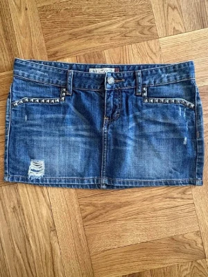 Blå jeanskjol - Snygg blå jeanskjol  med slitningar och coola nitar vid fickorna. Klassisk femficksmodell med bälteshällor och råa detaljer. Perfekt för dig som gillar en edgy och trendig stil.