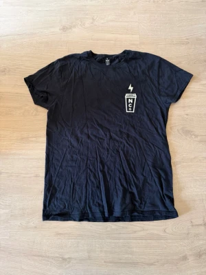 Svart t-shirt från Northern Coffee & Gear - Svart t-shirt med tryck fram och bak från Northern Coffee & Gear. Framsidan har en liten kopp med blixt och NCG-logga, baksidan har ett stort dödskalletryck och texten 'Death Before Decaf'. Skön passform och klassisk rund hals.