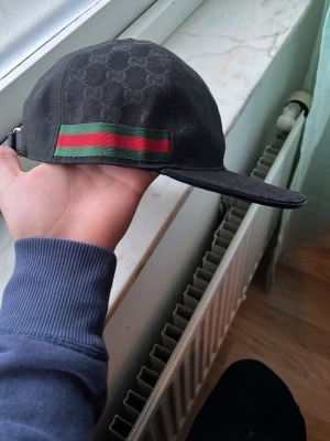 Svart Gucci keps med GG-mönster - Snygg svart keps från Gucci med klassiskt GG-monogram över hela kepsen. På sidan finns den ikoniska gröna och röda randen. Justerbar rem bak för perfekt passform. Perfekt accessoar för dig som vill ha en exklusiv och trendig look.