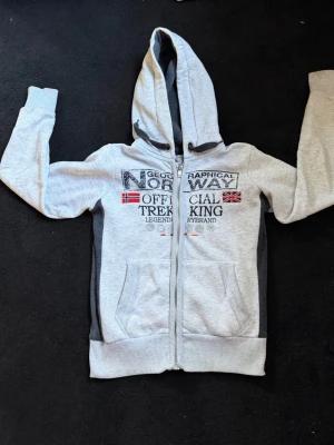 Grå hoodie från Geographical Norway - Snygg ljusgrå hoodie med dragkedja från Geographical Norway. Tryck på bröstet med norska och brittiska flaggan samt texten 'Official Trekking'. Mörkgrå paneler på sidorna, två fickor fram och justerbar huva. Perfekt för chill eller äventyr.