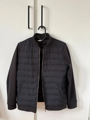 Svart cardigan Jacka - Snygg svart cardiganjacka med quiltad framsida och baksida, ribbade muddar och krage. Jackan har dragkedja framtill och två fickor med dragkedja. Ärmarna är i ett mjukare tyg som kontrasterar mot den glansiga kroppen. Perfekt för kalla vinterdagar.