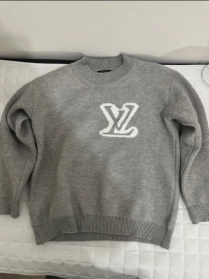 Grå Louis Vuitton tröja med logga - Snygg grå stickad tröja från Louis Vuitton med vit LV-logga framtill. Tröjan har rund halsringning, ribbade muddar och är tillverkad i mjukt material som passar perfekt för kyligare dagar.