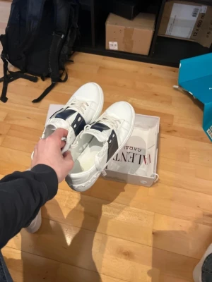 Valentino Garavani skor - Snygga vita sneakers från Valentino Garavani med bred mörkblå rem över mitten och coola nitar på sidan. Skorna har en klassisk låg profil och är tillverkade i slätt läder med vita snören och sula. Perfekta för dig som gillar exklusiv och stilren design.