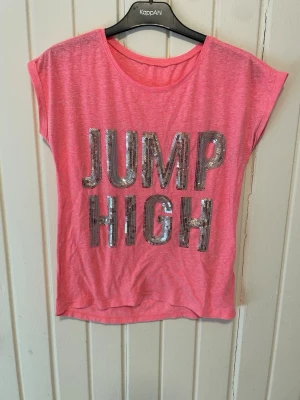 Rosa t-shirt med paljetter från H&M - Cool rosa t-shirt från KappAhl med texten 'JUMP HIGH' i silvriga paljetter framtill. T-shirten har korta ärmar och en avslappnad passform. Perfekt för dig som gillar att sticka ut med lite extra glitter i vardagen.